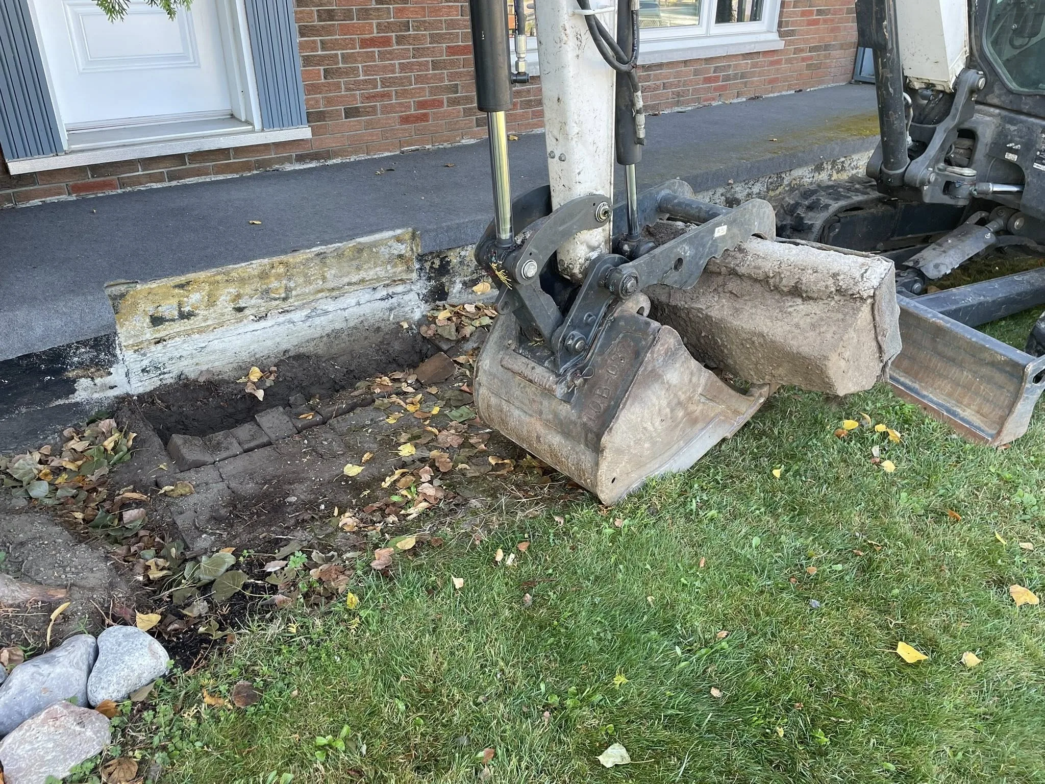 Mini excavator demolishing a failing concrete walkway during a Bouwers Design Landscaping driveway replacement in North Bay, Ontario.
