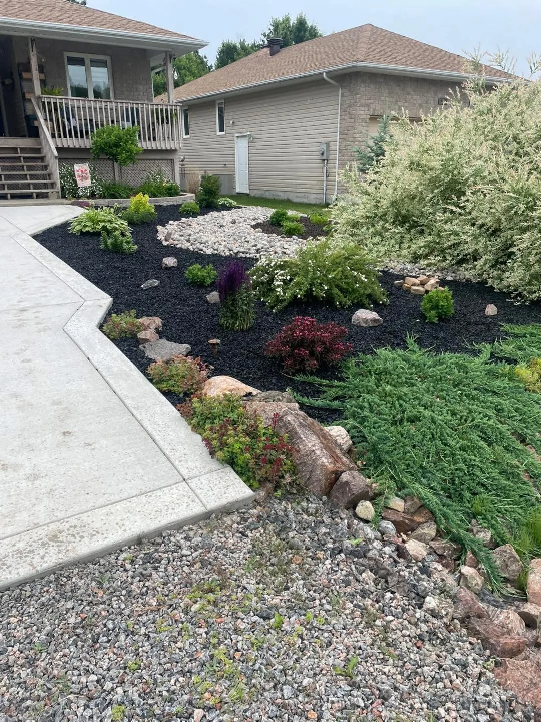 Finished Bouwers Design Landscaping property in North Bay, Ontario, with mature plantings, a decorative river-rock dry creek, mulched beds, and feature stones integrated against the home.