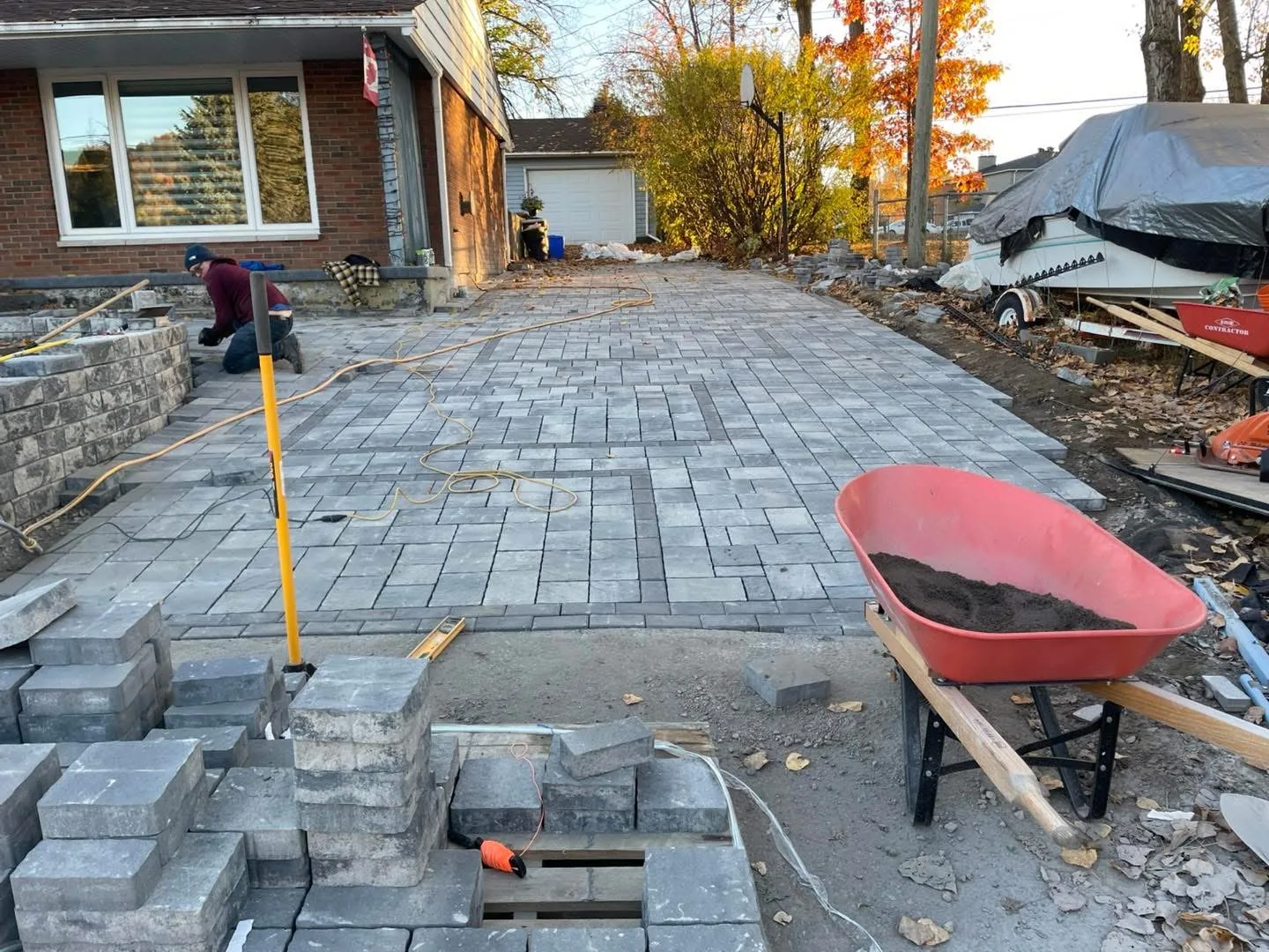 Bouwers Design Landscaping crew member hand-laying a Permacon paver driveway in North Bay during an autumn install, with a polymeric sand wheelbarrow staged at the working edge.