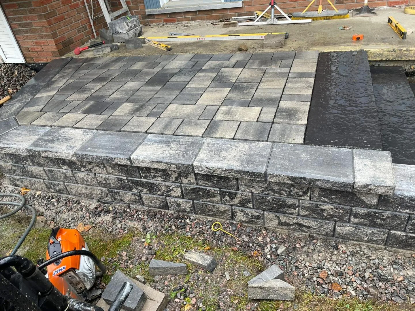 Permacon block retaining wall with cap course and integrated paver landing on a Bouwers Design Landscaping porch build in North Bay, Ontario, photographed mid-build with the Stihl saw set down.